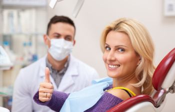 A dentist and a satisfied woman with perfect smile sitting in a dental chair and showing her thumb up.